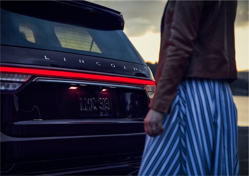 A person is shown near the rear of a 2023 Lincoln Aviator® SUV as the Lincoln Embrace illuminates the rear lights | Pines Lincoln in Pembroke Pines FL
